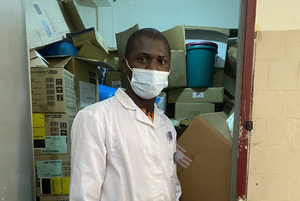 Dr Gabriel GOEPOGUI, Service pédiatrie-Hôpital Ignace Deen, point focal du projet Isanco devant le stock de matériel d'hygiène et de protection fournis dans le cadre du projet Isanco pour mieux protéger le personnel de santé et les patient·es qui fréquentent le service.